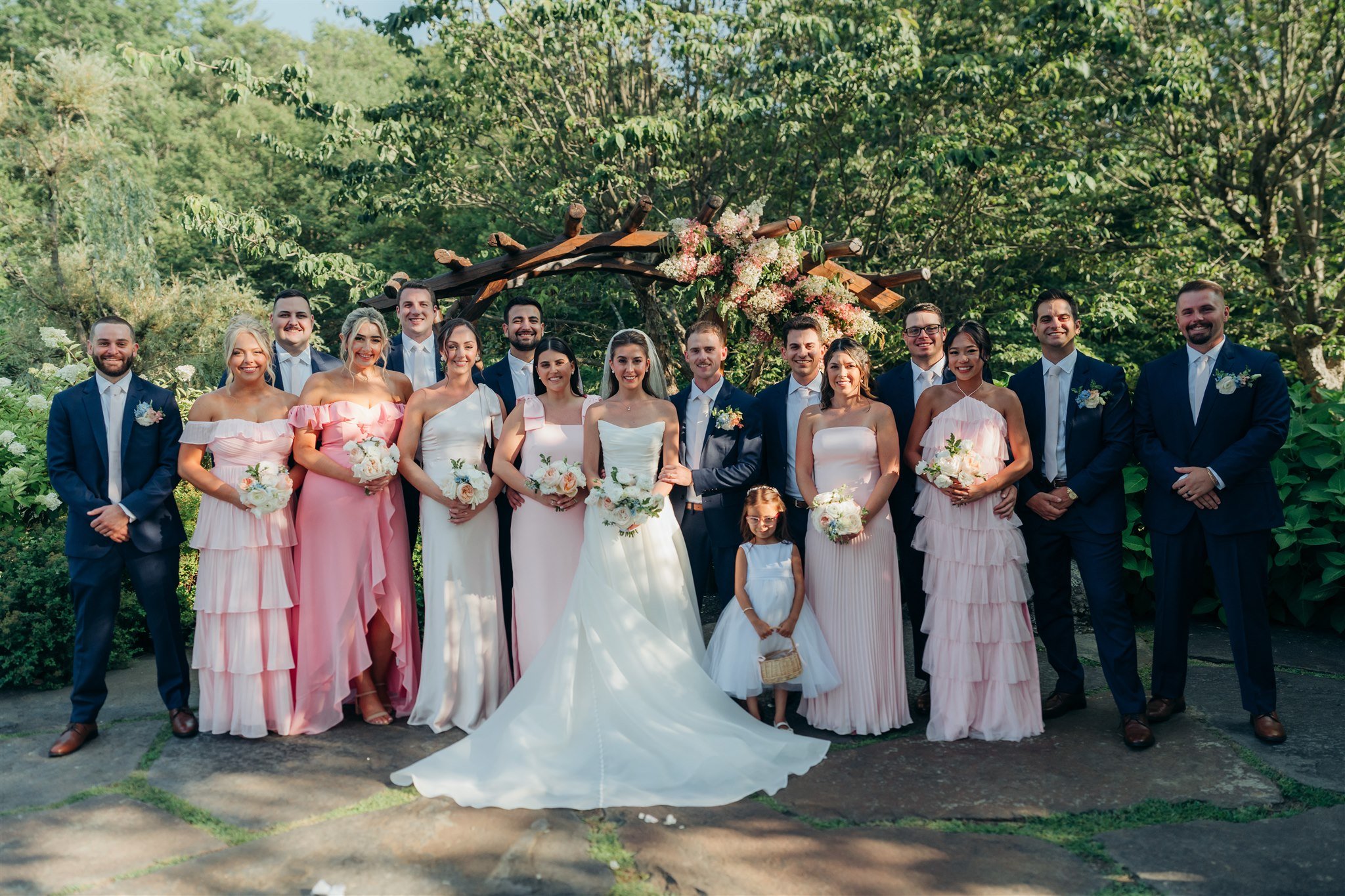 A group of wedding party members posing outdoors under a floral arch with green trees in the background. Bridesmaids wear pink dresses, groomsmen wear navy suits with white shirts, and the bride and groom stand in the center with the bride in a white