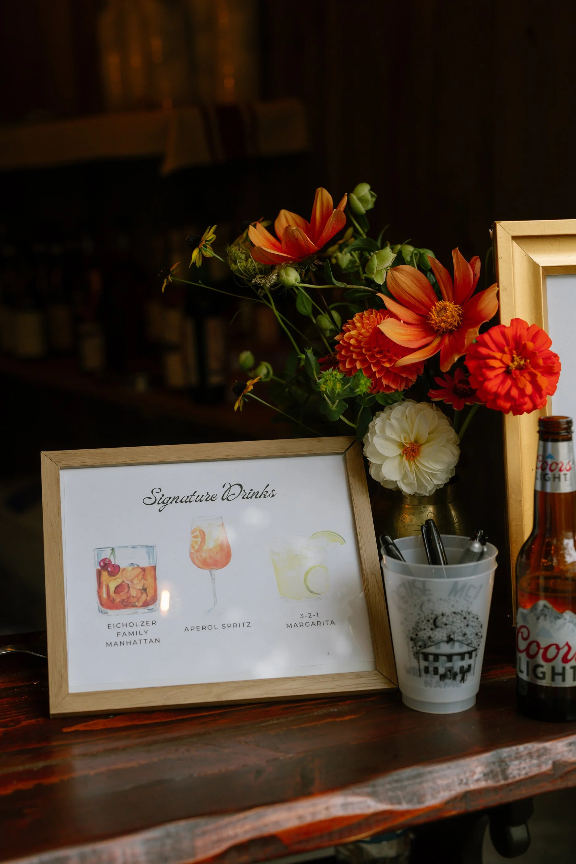A framed sign displaying three signature drinks, a bouquet of orange and white flowers in a vase, a container with pens, and a Coors Light beer bottle on a wooden counter.