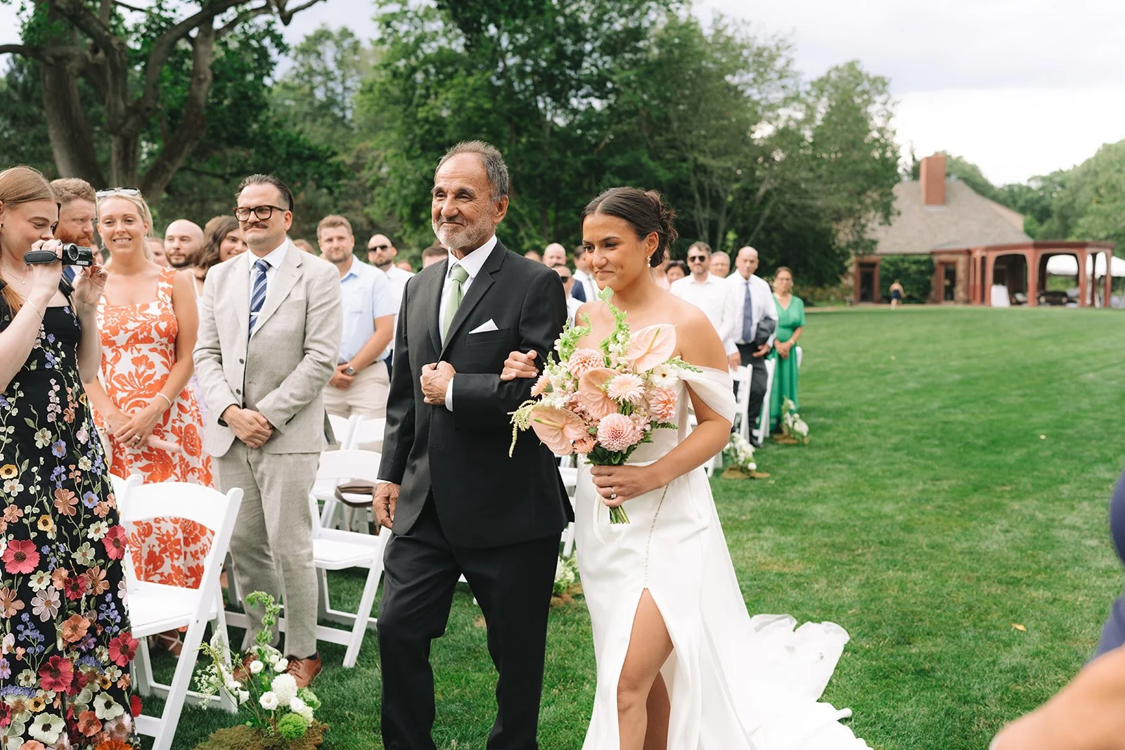 A bride walking down the aisle with her father at an outdoor wedding ceremony, holding a bouquet of pink and white flowers, with guests standing in the background.