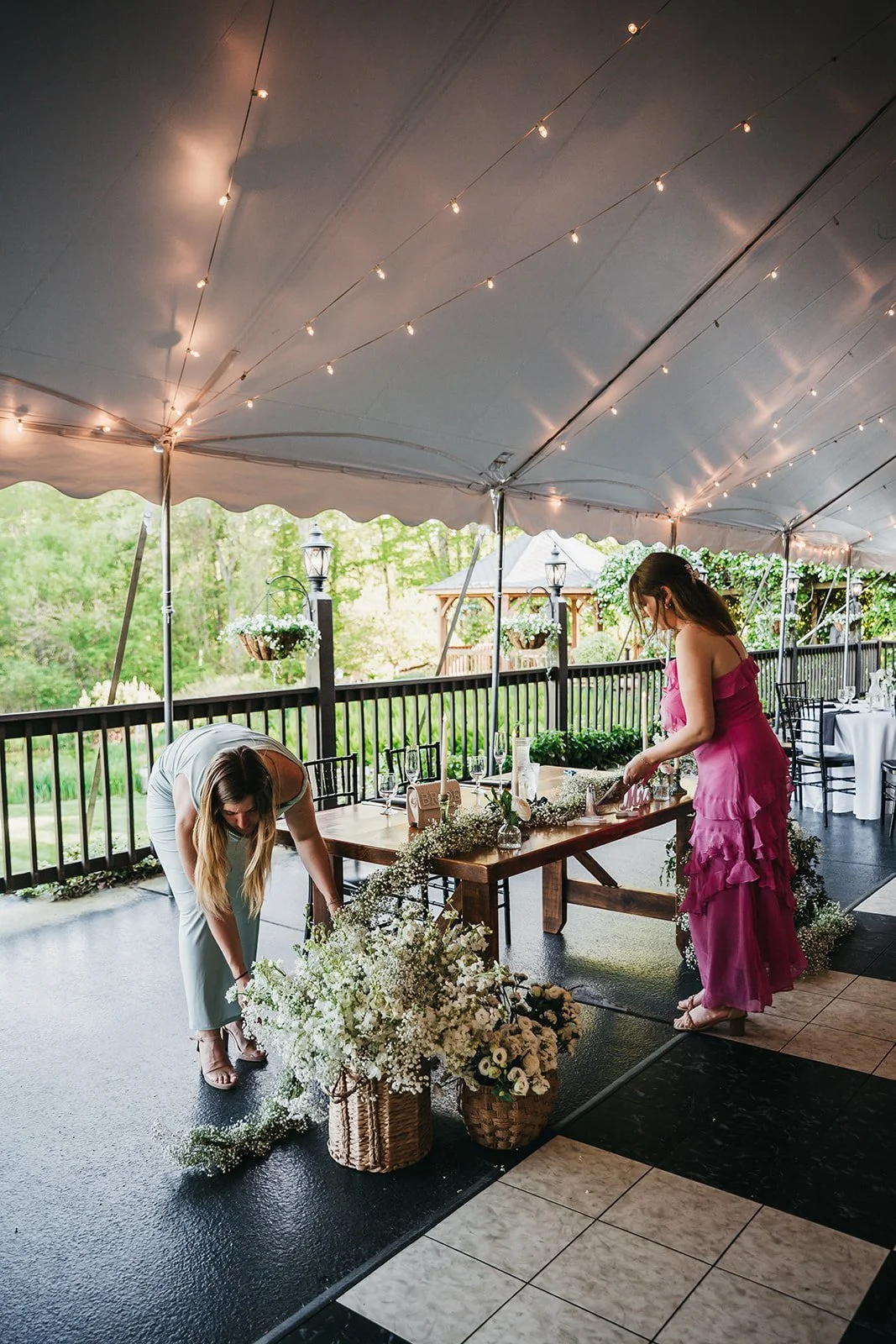 Two women preparing floral arrangements at a decorated outdoor wedding reception under a large tent with string lights.