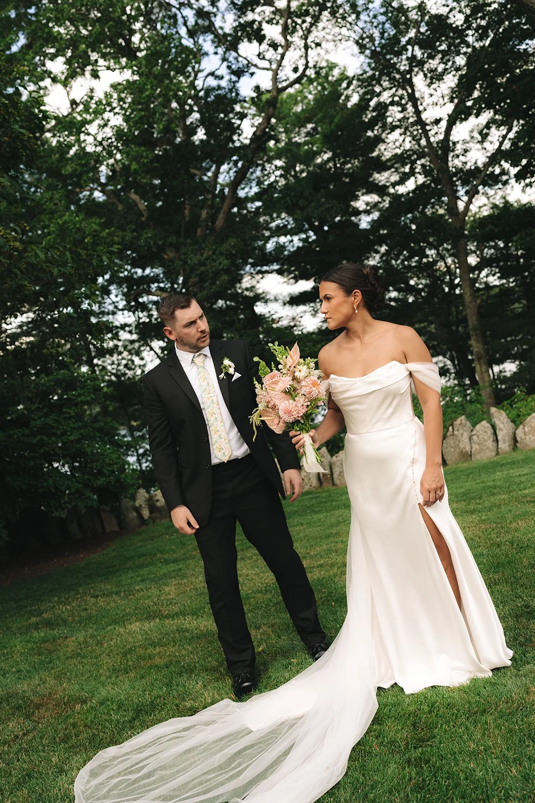 A bride in a white off-shoulder wedding gown with a slit and train holds a bouquet of pink and white flowers, standing on a grassy area surrounded by trees, while a groom in a black suit with a floral tie looks on.