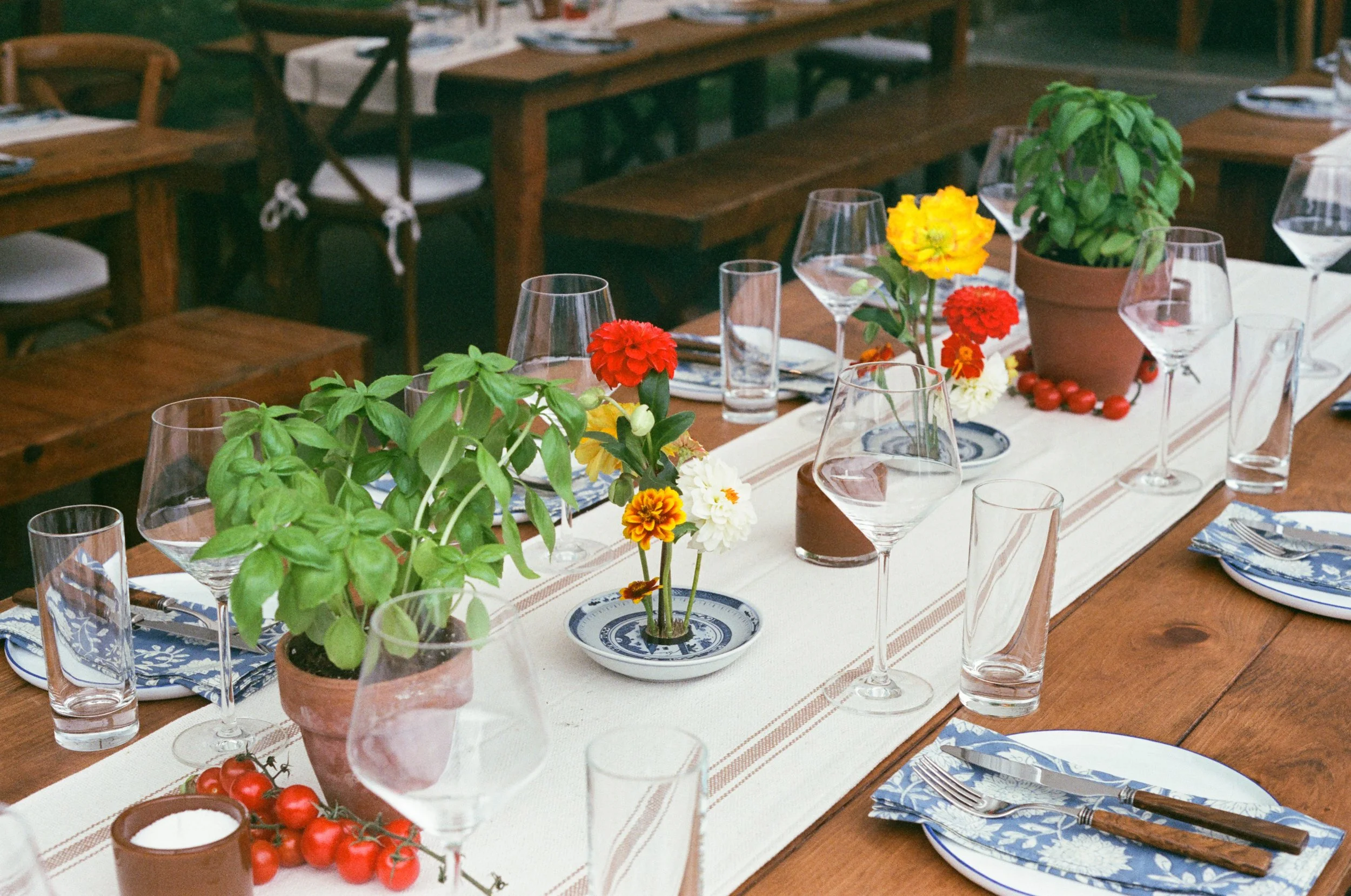 A long wooden dining table set for a meal with floral centerpieces, wine glasses, plates, and cutlery, and additional wooden tables and chairs in the background.