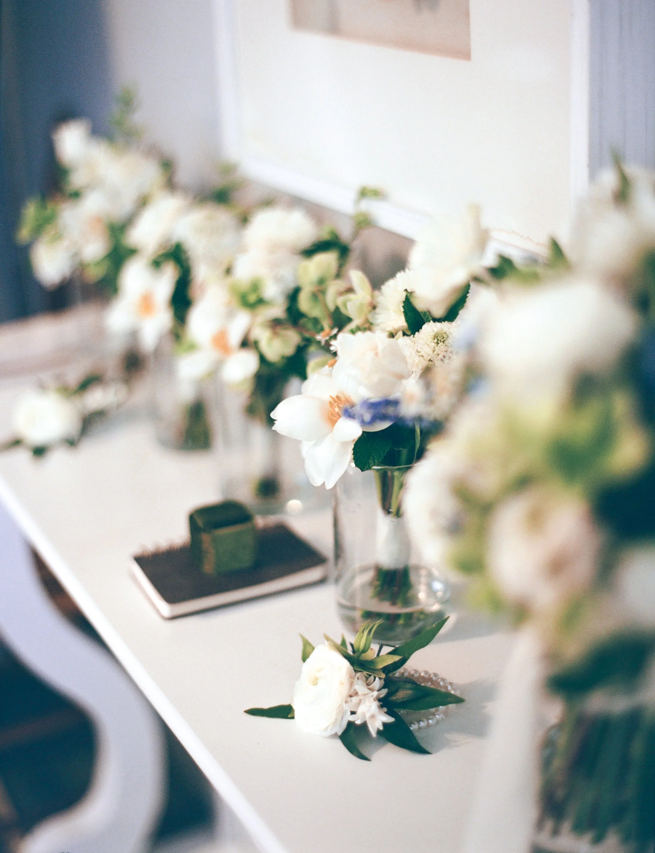 A row of white flower bouquets in glass vases on a white table, with a notepad and green box in the background.
