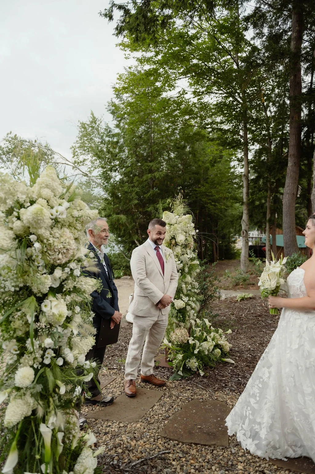 A wedding ceremony outdoors with a bride in a white lace gown holding a bouquet, standing near a groom in a beige suit, facing each other, with an officiant and a man standing behind floral arrangements and green trees in the background.