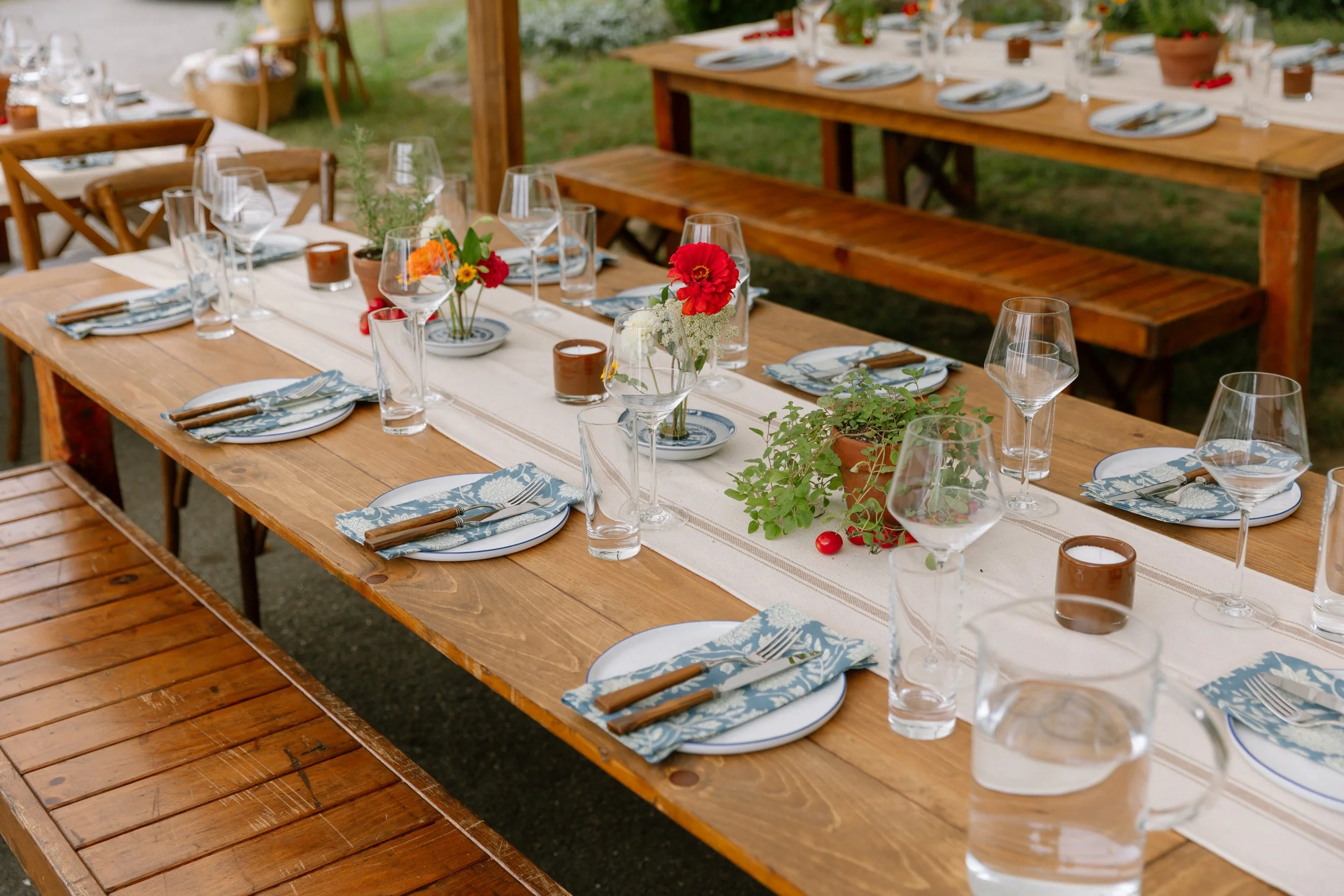 A long wooden outdoor dining table set for a meal with plates, silverware, glasses, flower arrangements, and candles, on a grassy area.
