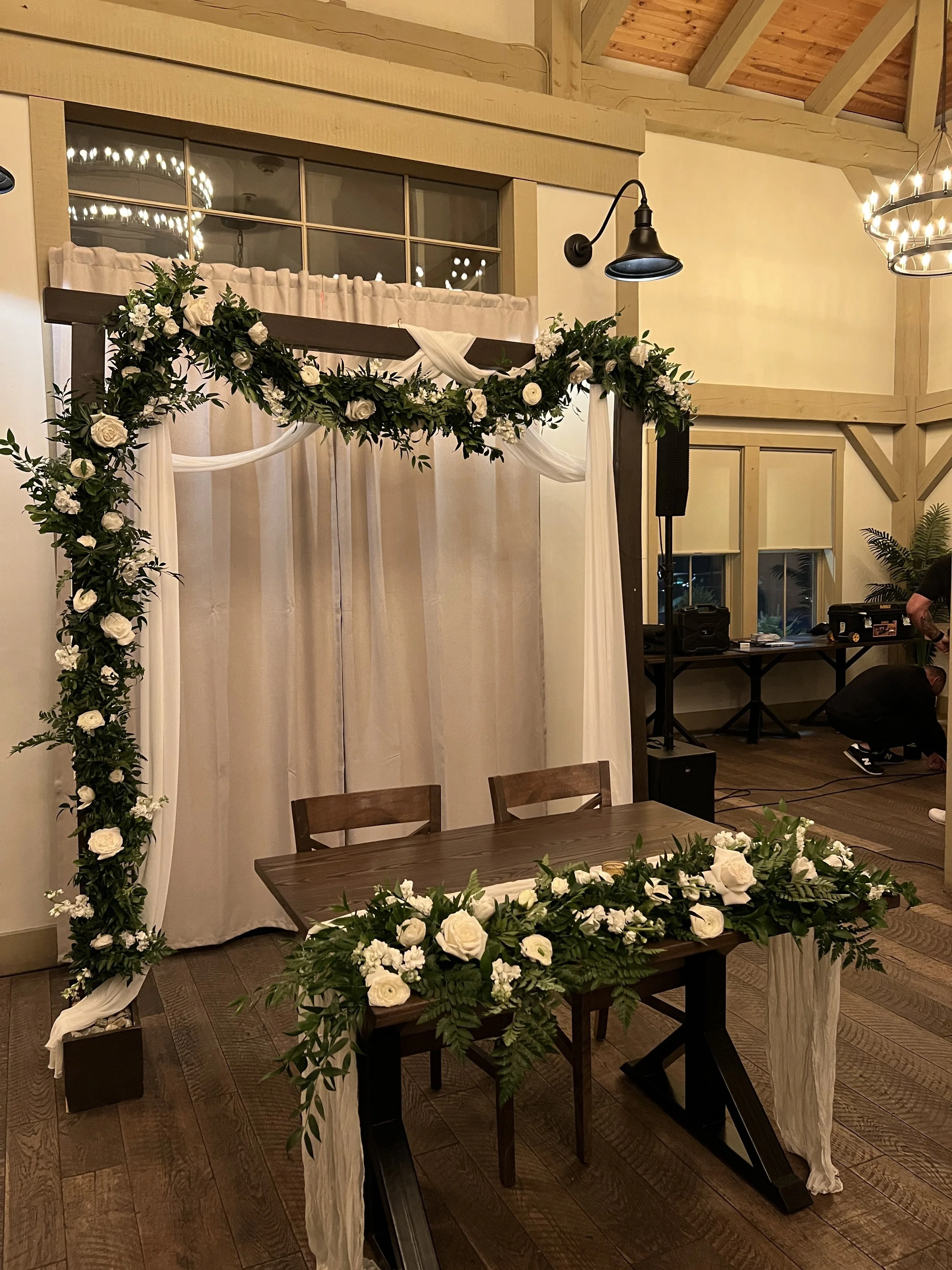 Wedding altar with white roses and greenery, wooden table with matching floral arrangement, behind curtains in a rustic indoor venue.