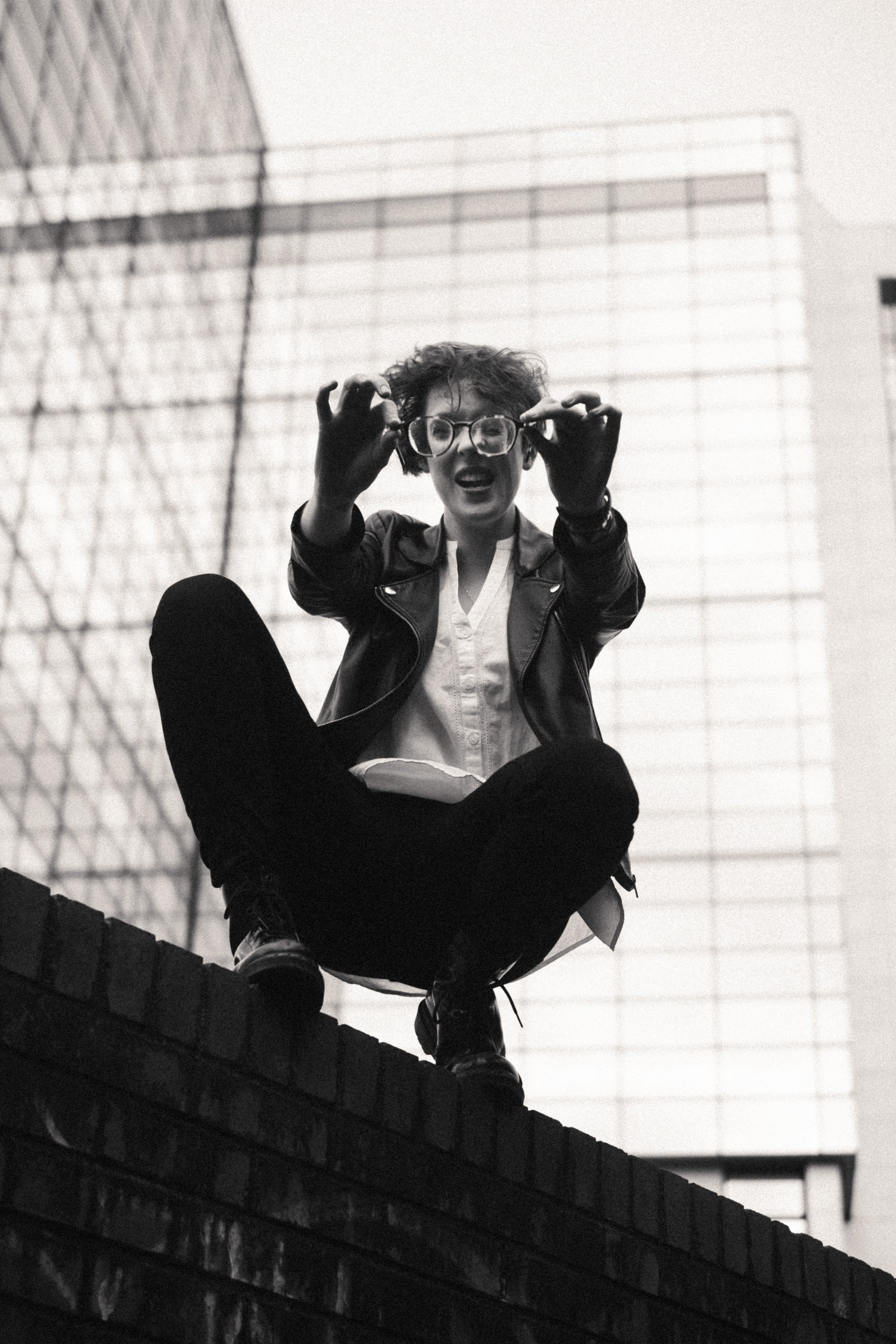 Person sitting on a brick wall, wearing glasses and a leather jacket, in front of a glass building, black and white photograph.