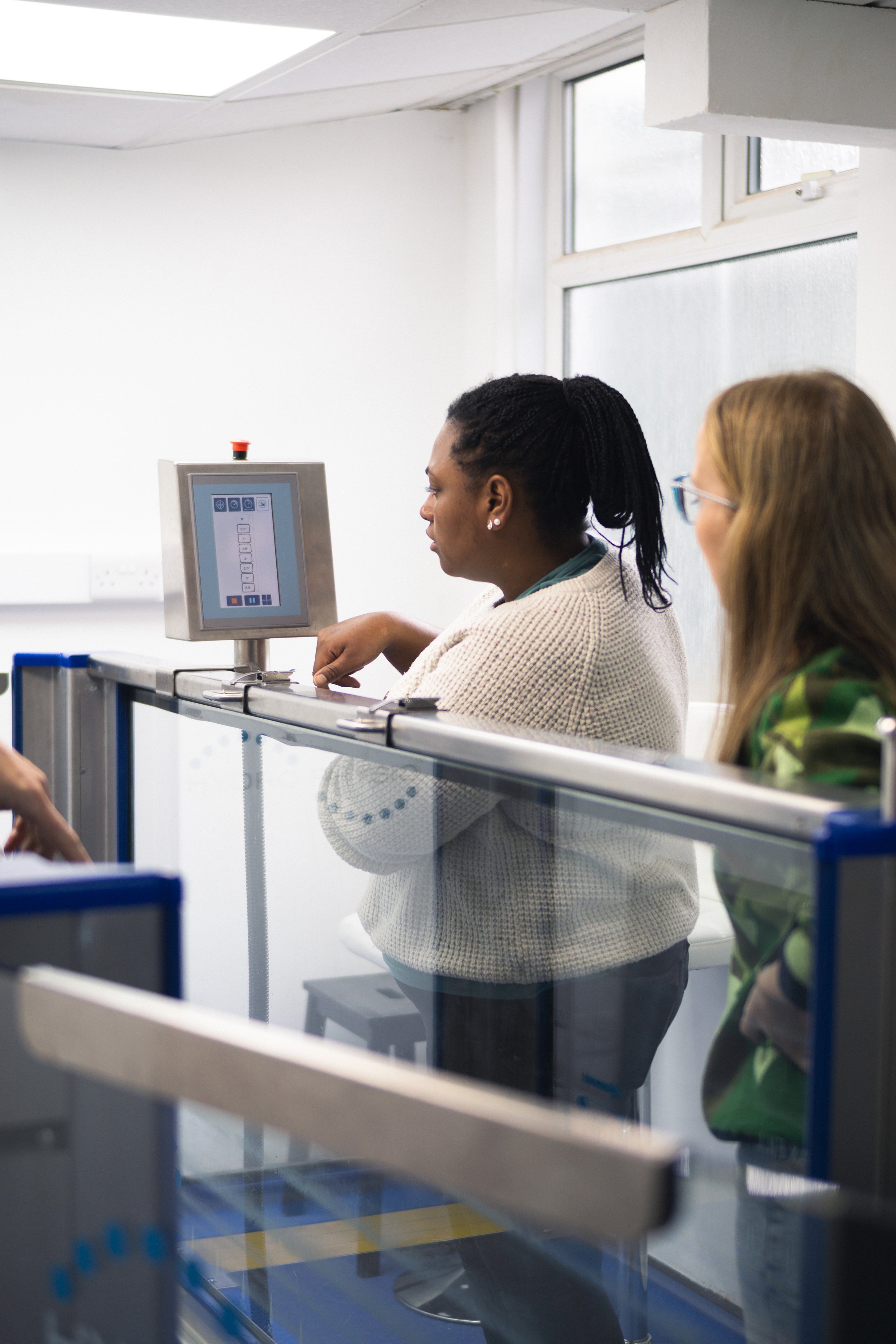 Two women standing inside a room, one using a touchscreen interface, with a glass partition in front.