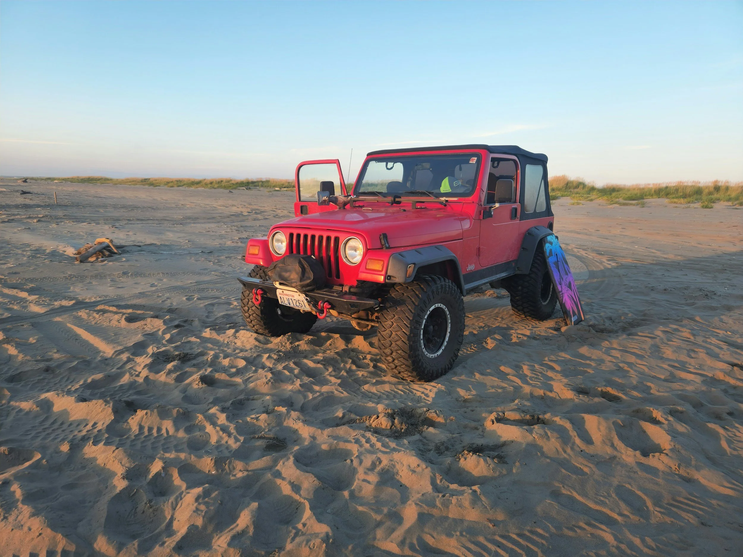 Red Jeep parked on sandy beach with a purple and black skateboard leaning against the tire, beach grass in the background, and a clear blue sky.