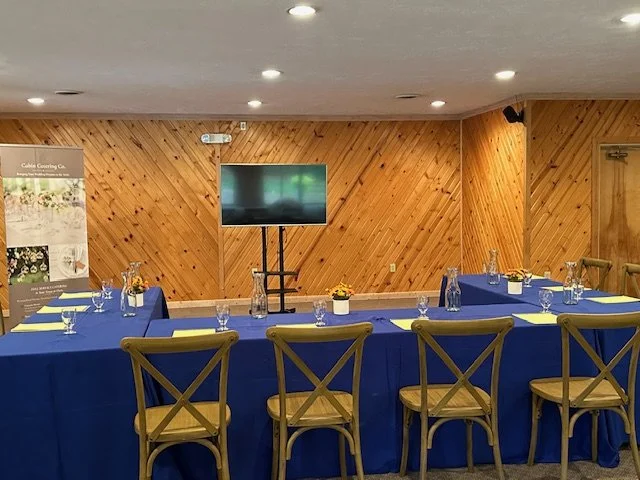 Corporate meeting room with a U-shaped table covered in blue tablecloths, set with glasses, notebooks, and flowers, a flat-screen TV on a stand, wood-paneled walls, and chairs placed around the table.