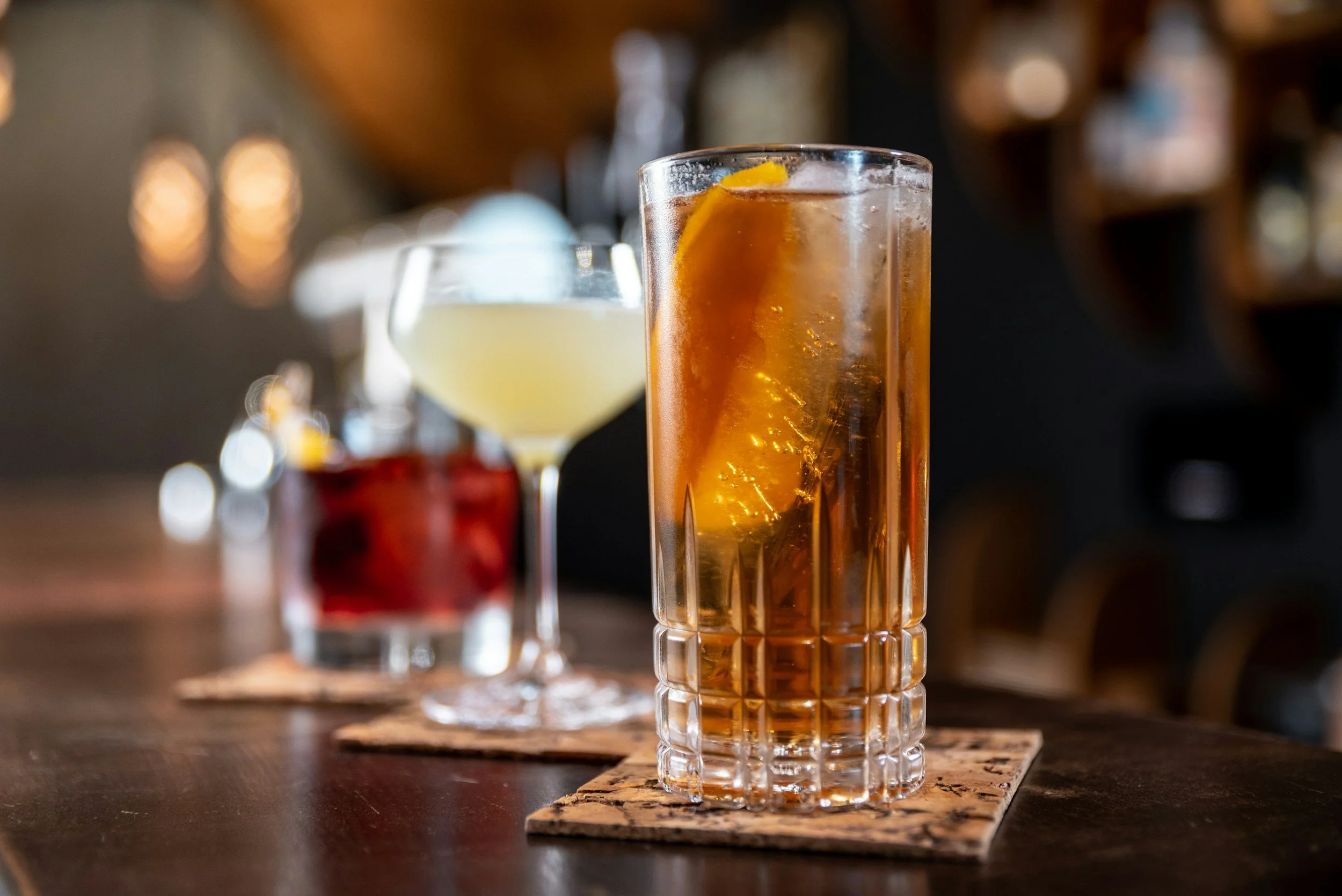Three cocktails on a bar counter with a blurred background. The front drink is a tall glass with ice and a lemon wedge, the middle drink is a coupe glass with a light-colored cocktail, and the back drink is a short glass with a dark red beverage and a lemon garnish.