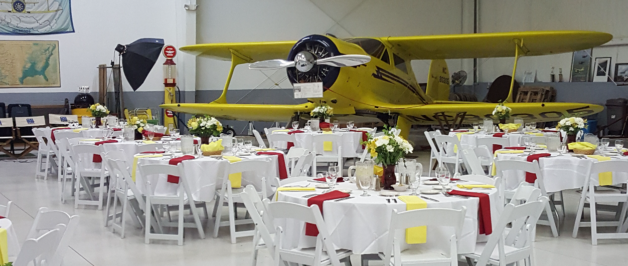 A vintage yellow airplane displayed behind a banquet-style dining event setup with round tables, white tablecloths, and red and yellow napkins, in an indoor airplane hanger setting.