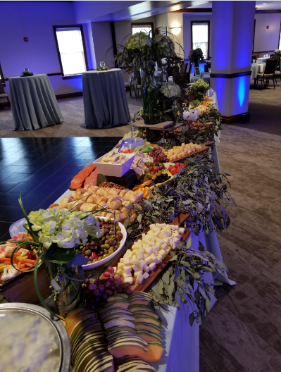 A grazing table set with charcuterie of cheese, meats, grapes, crackers, and fruit at an indoor event venue with blue lighting.