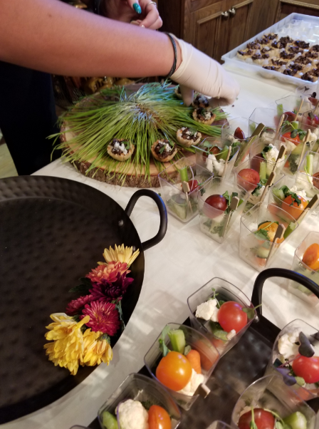 Person preparing finger foods, including small desserts and vegetable cups, on a table with black trays and floral decorations.