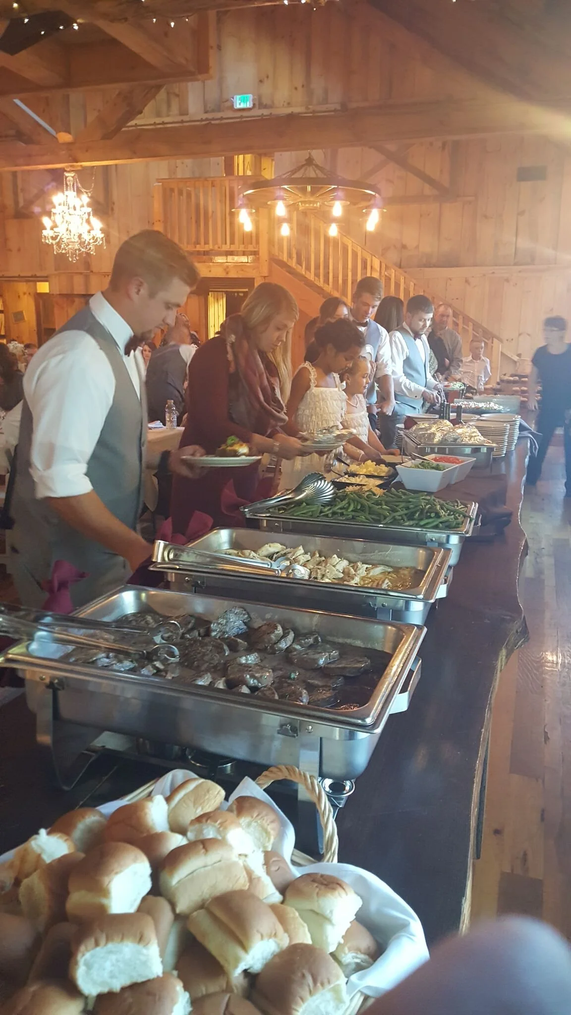 People serving themselves food at a buffet in a rustic wooden venue with chandeliers and warm lighting.