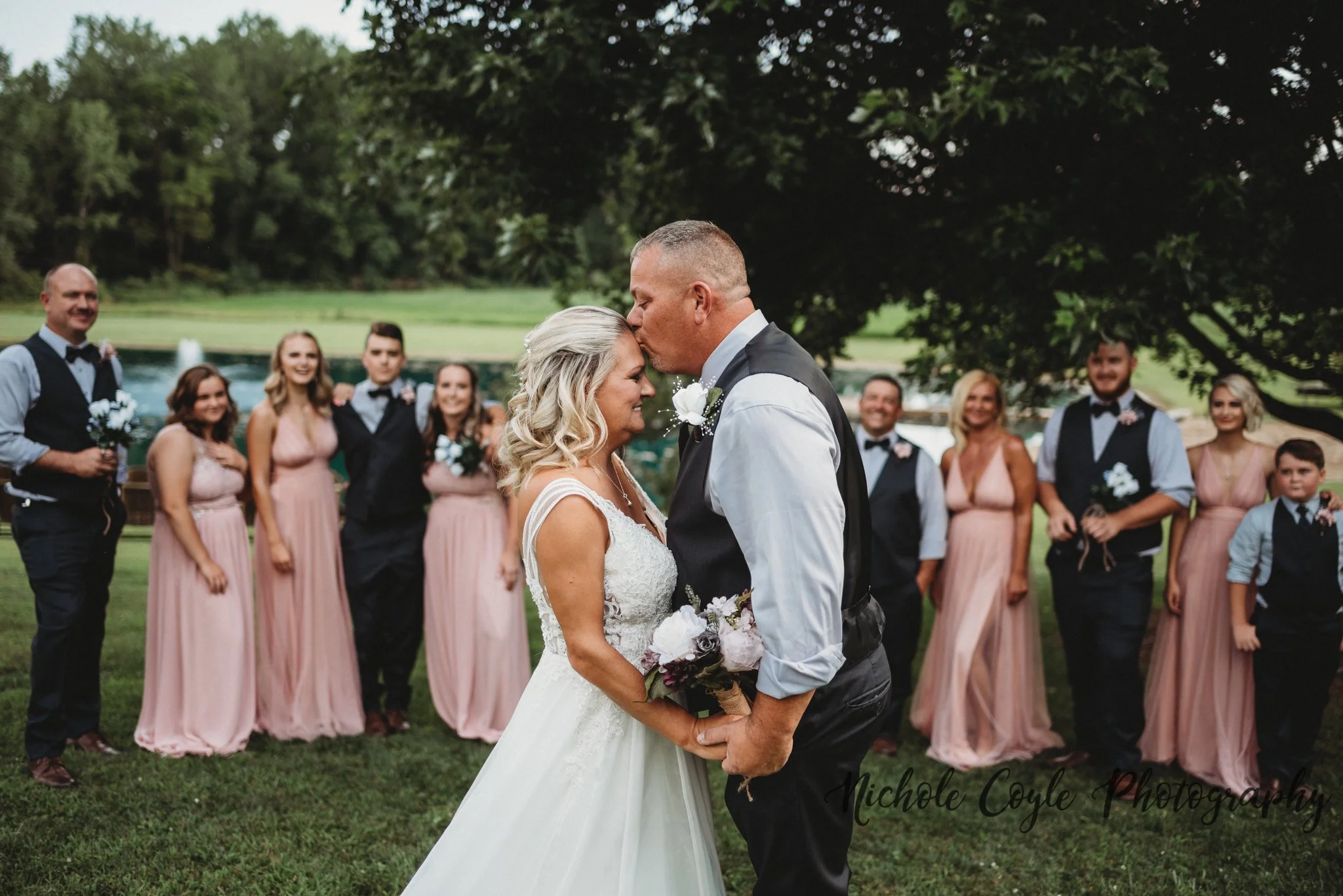 A bride and groom share a kiss during their wedding ceremony outdoors, surrounded by bridesmaids and groomsmen, in a park with trees and a fountain in the background.