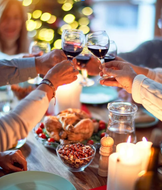 People celebrating with glasses of red wine at a festive dinner table with a roasted turkey, candles, and holiday decorations.