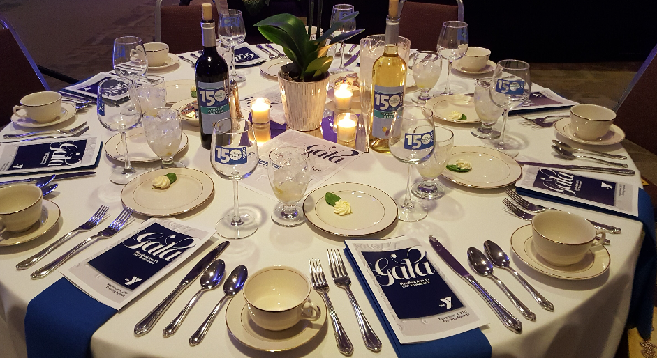 A round table set for a corporatgala event with plates, cutlery, wine glasses, and cups. A center potted plant and lit candles decorate the table. Bottles of wine are also present. Program guides with 'Gala' written on them are at each place setting.