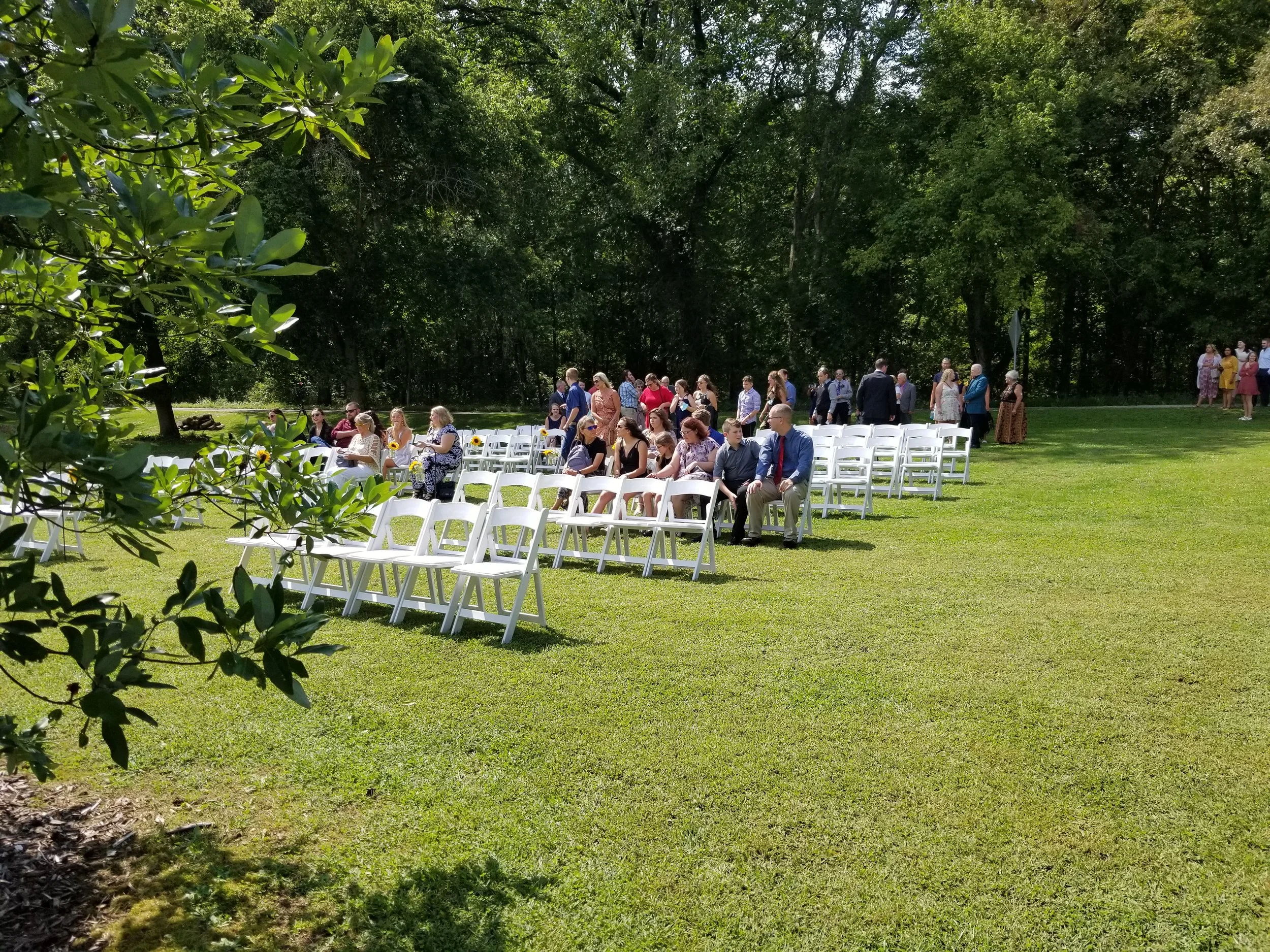 People attending an outdoor event or wedding, seated on white chairs on a grassy lawn surrounded by trees.