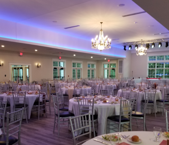Wedding reception venue with round tables, white tablecloths, and silver chairs, decorated with pink napkins and plates, illuminated by chandeliers and blue LED lighting.
