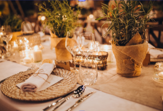Elegant dining table setup with woven placemats, white napkins, wine glasses, candles, and potted plants wrapped in burlap.