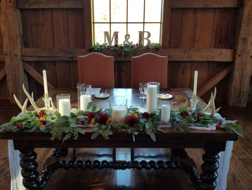 A rustic wedding or event table decorated with candles, greenery, and flowers, with large antlers on either side, two chairs at the back, and a window with the letters M and B in the background.