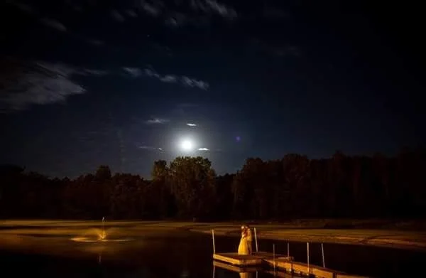 Night scene of a Bride and Groom sitting on a dock by a lake, with a bright moon and stars in the sky; trees are visible in the background at a rural wedding venue.