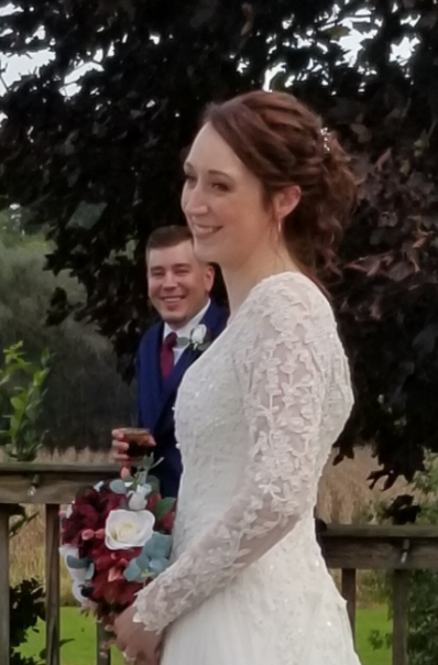 A bride and groom standing outdoors with trees in the background. The bride is in a white lace wedding dress and the groom is in a blue suit holding a bouquet.