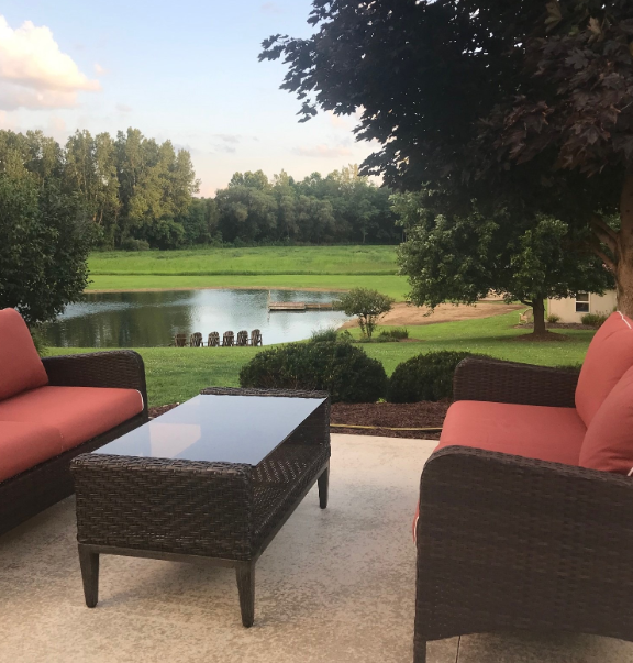 Outdoor patio with wicker chairs and a glass-topped table overlooking a pond and lush greenery.