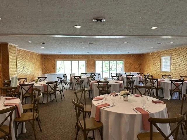 Round dining tables with white tablecloths, pink napkins, and glassware, arranged in a banquet-style in a decorated rustic event venue with wood-paneled walls.