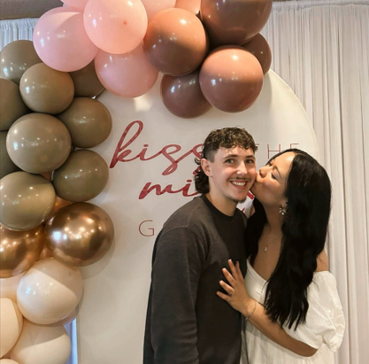 A young woman kissing a young man on the cheek at a bridal shower with a balloon arch backdrop that says "kiss me."