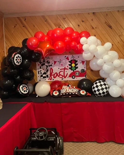 Balloon garland with red, black, white, and checkered balloons, car-themed balloons, and a birthday banner with racing and car motifs, set on a wooden-paneled wall backdrop.