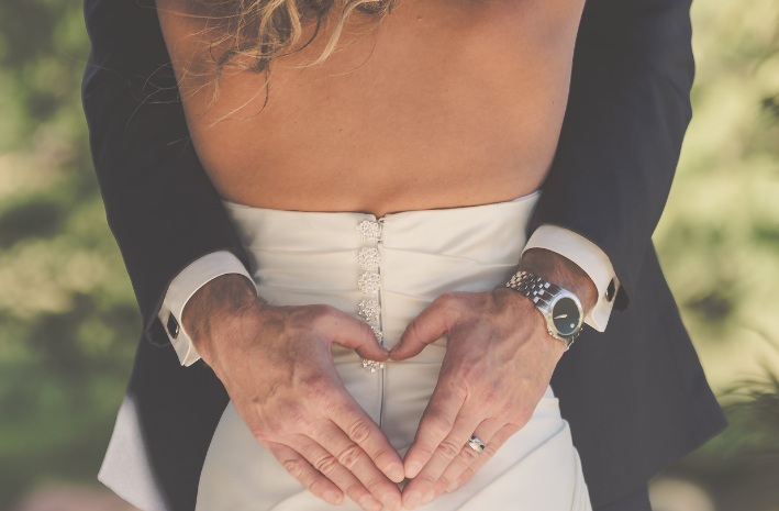 A close-up of a bride and groom's hands forming a heart shape during a wedding photo, with the bride wearing a white gown and the groom in a black suit.