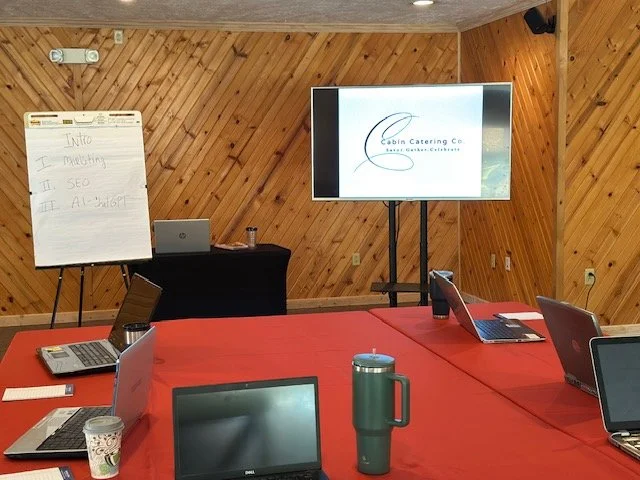 A corporate conference room with a red tablecloth-covered table, multiple laptops, a flip chart on a stand, a large mounted screen displaying a logo for Cabin Catering Co., and a black table with a laptop and a can on it.