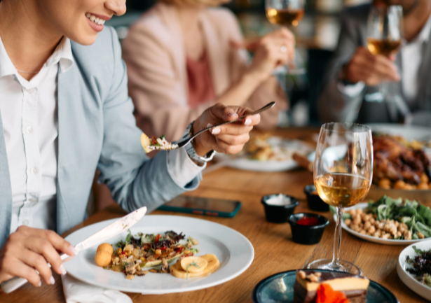 People dining at a restaurant, enjoying food and drinks, with a woman smiling and eating salad, others holding glasses of wine, and various dishes on the table.