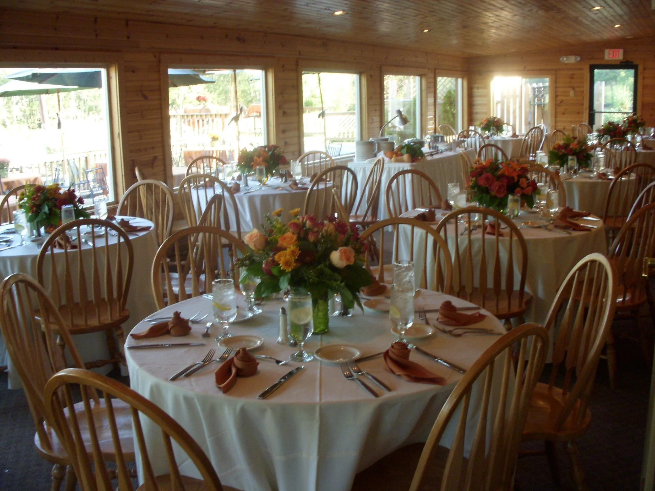 Elegant banquet hall with round tables covered in white tablecloths, set with glassware, silverware, fall accents and floral centerpieces, with rustic wooden walls and large windows allowing natural light.