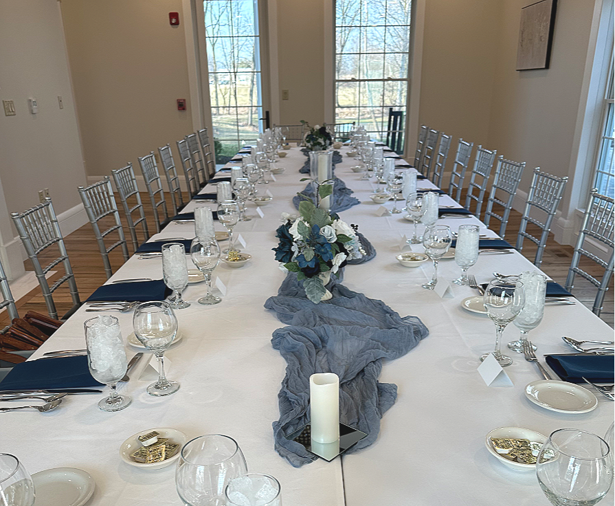 Long dining table set for a corporate formal event with white tablecloth, floral centerpiece, blue napkins, and glassware, in a room with large windows.