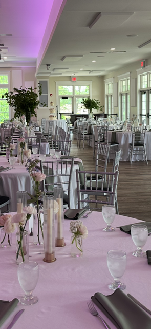 Elegant event venue set up for a wedding or event with round tables covered in pink tablecloths, silver chairs, floral centerpieces, candles, and glassware, with natural light coming through large windows.