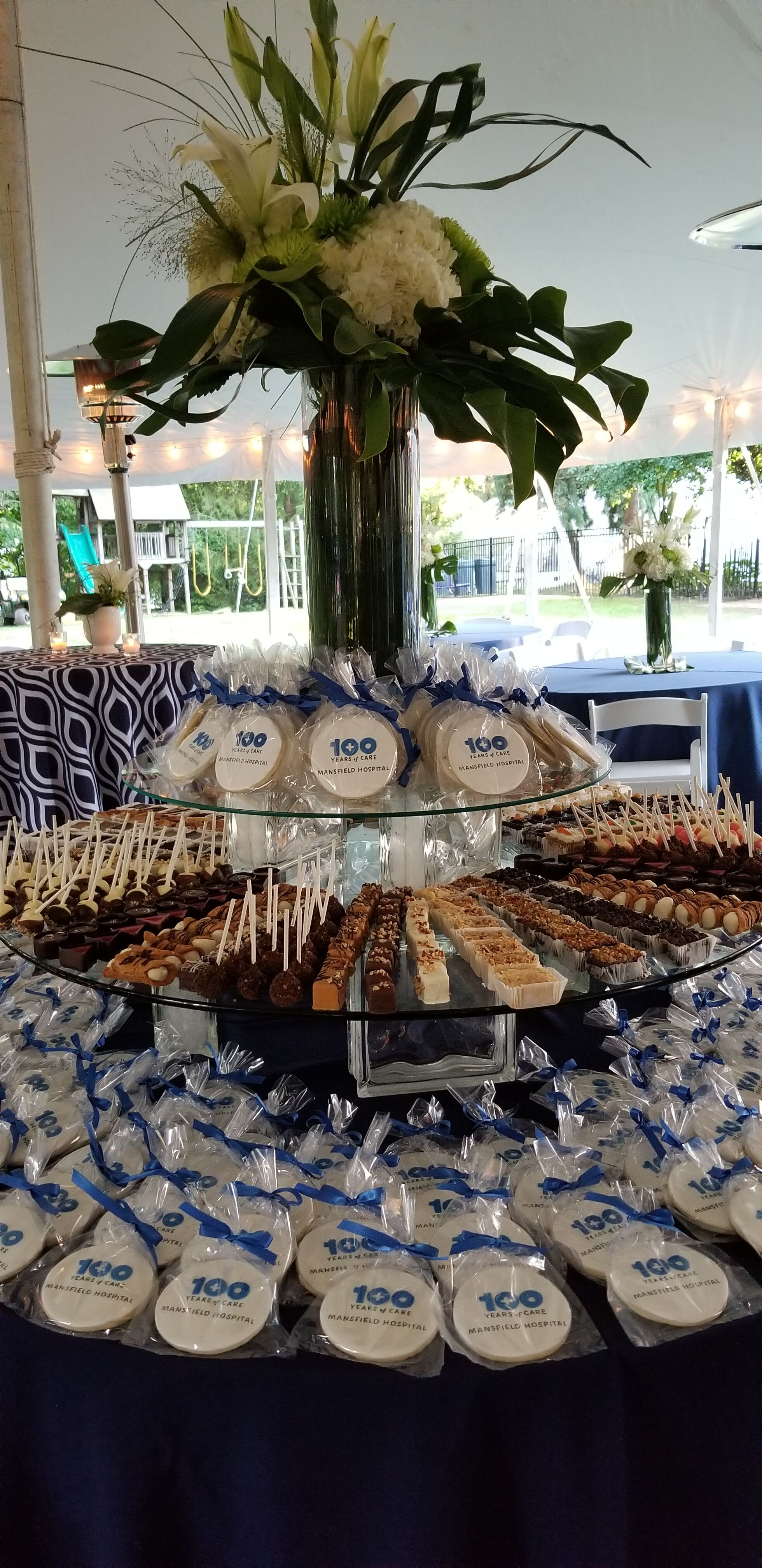 A dessert table with an assortment of sweets and cookies with a large floral arrangement in a tall vase, 100th anniversary celebration of care at Ohio Health Mansfield Hospital.