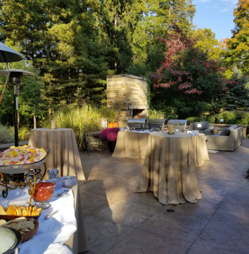 Outdoor party setup with tables, chairs, a food table, and a brick fireplace surrounded by trees and greenery.