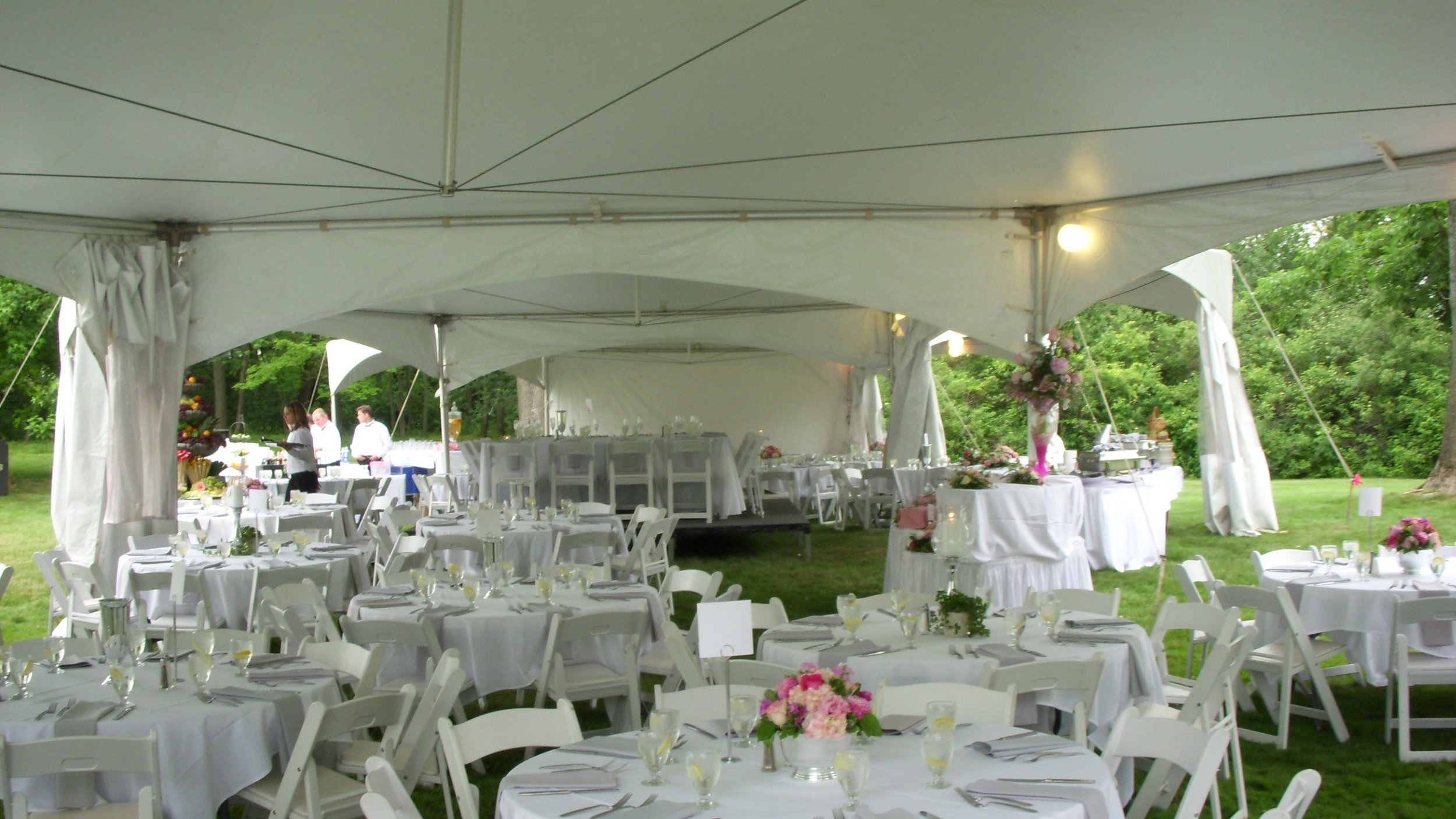 Outdoor wedding reception setup under a large white tent with round tables, white chairs, floral centerpieces, and buffet tables, surrounded by green trees