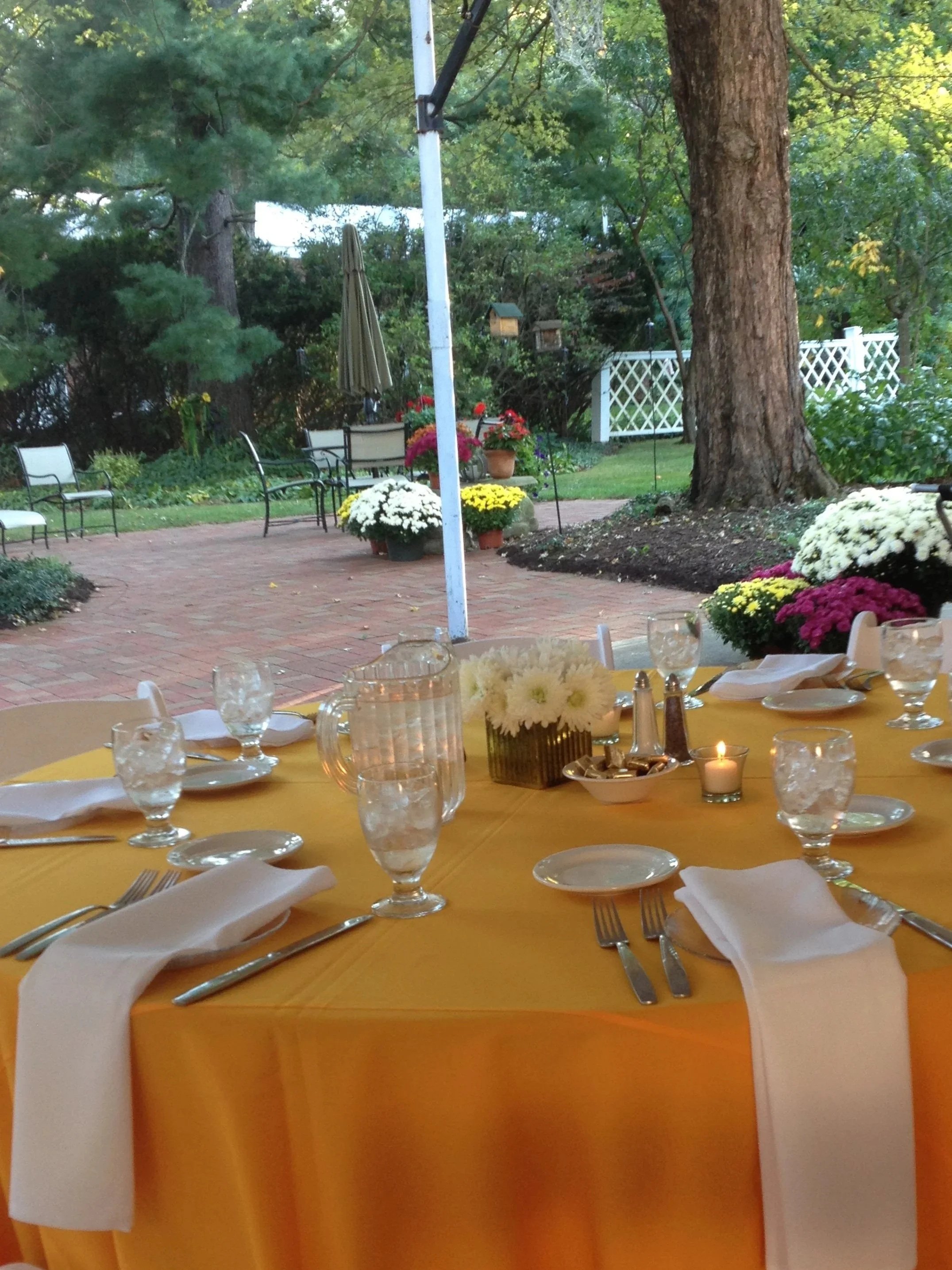 Decorated outdoor rehearsal event with a yellow tablecloth, napkins, glasses, and silverware, set in a garden with potted flowers, trees, and a brick patio.