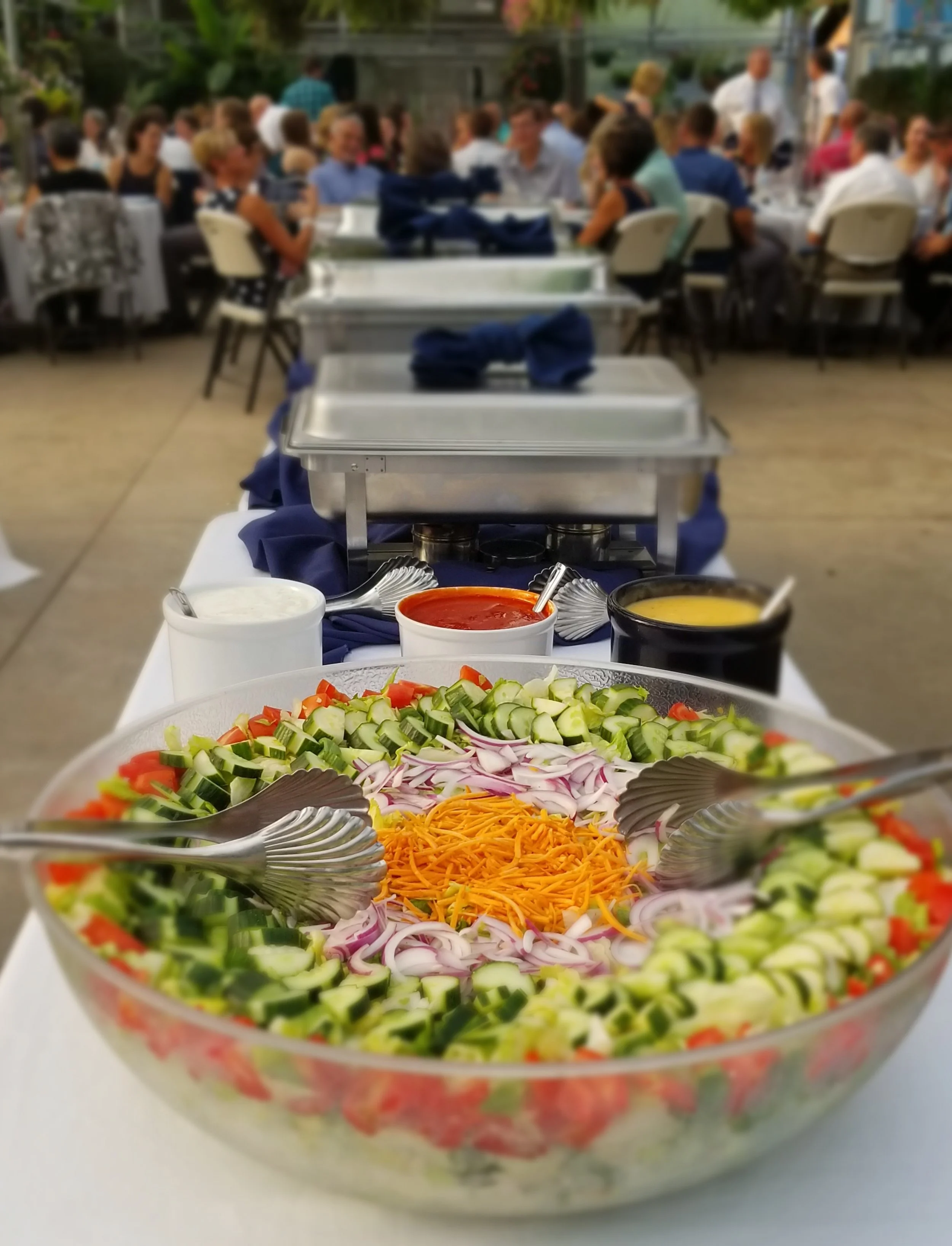 Large bowl of fresh salad with cucumbers, tomatoes, onions, and shredded cheese on a buffet table at a greenhouse event venue with people dining in the background.