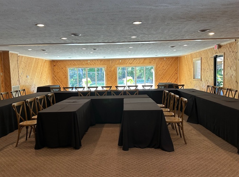 Corporate conference room with U-shaped tables covered in black tablecloths and wooden chairs around them for corporate meeting, large windows with greenery outside, wood-paneled walls.