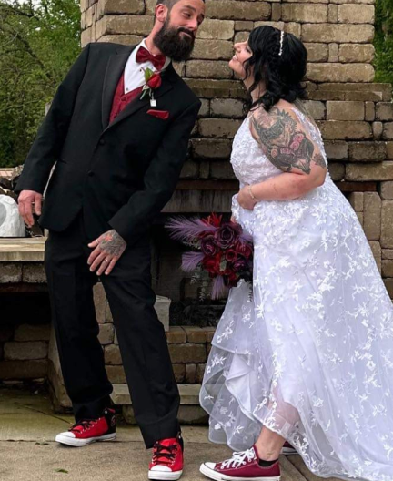 A bride and groom with contrasting styles, the bride in a white lace wedding dress holding a bouquet with red and purple flowers, and the groom in a black suit with a red shirt and sneakers, posing playfully in front of a brick wall.