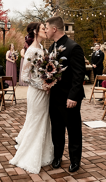 Bride and groom kissing during their outdoor wedding ceremony, with bridesmaid and groomsman in the background, decorative string lights overhead, brick floor, wooden chairs, and autumn trees.