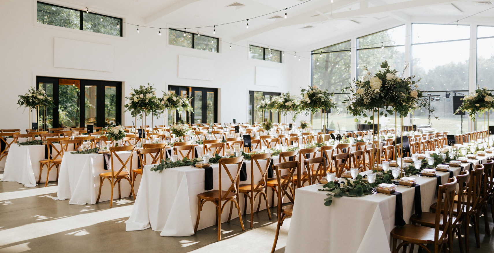 Elegant wedding reception setup in a bright, modern venue with large windows, decorated with white flowers and greenery, wooden chairs, and white tablecloths.