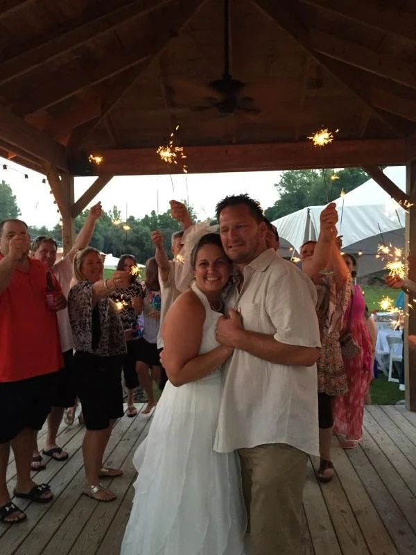 A couple dancing under a wooden pavilion at a celebration, with friends holding sparklers in the background.