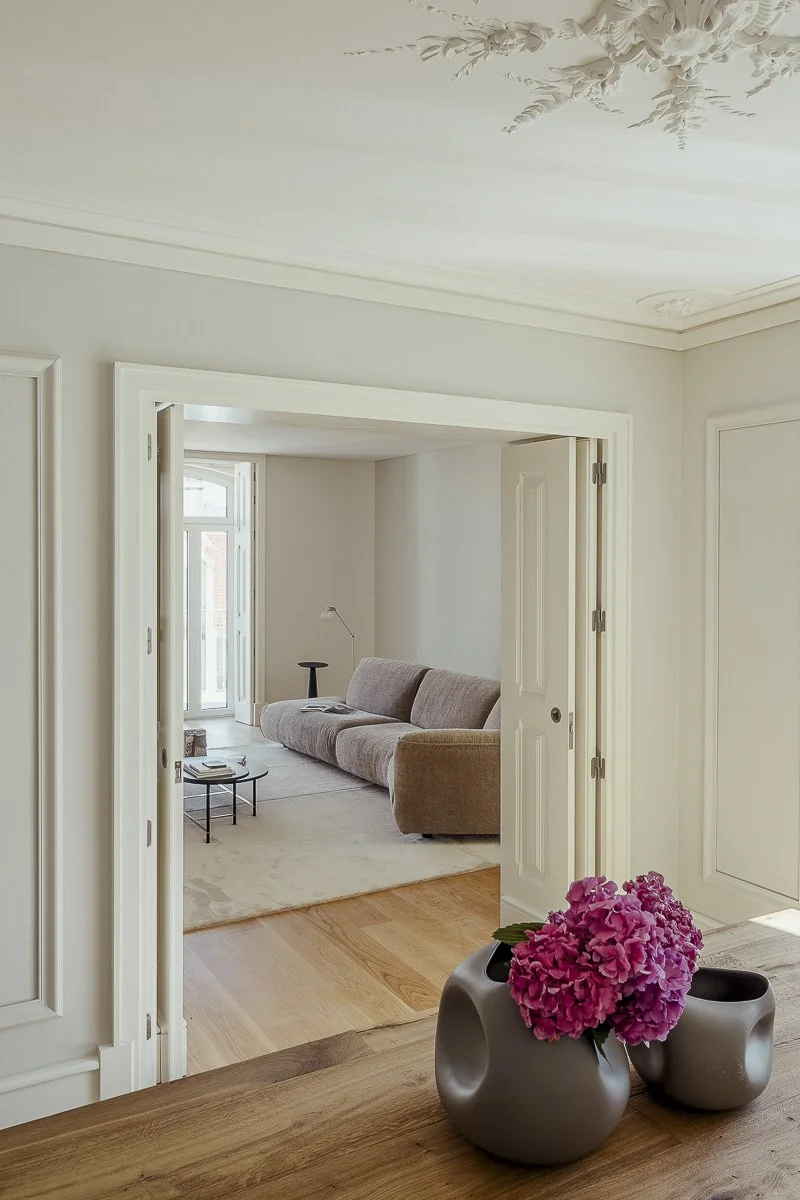 A view of a living room seen through an open doorway, with a beige sofa against the far wall, a small side table, and a window letting in natural light. In the foreground, on a wooden surface, there are two gray vases with pink hydrangea flowers.