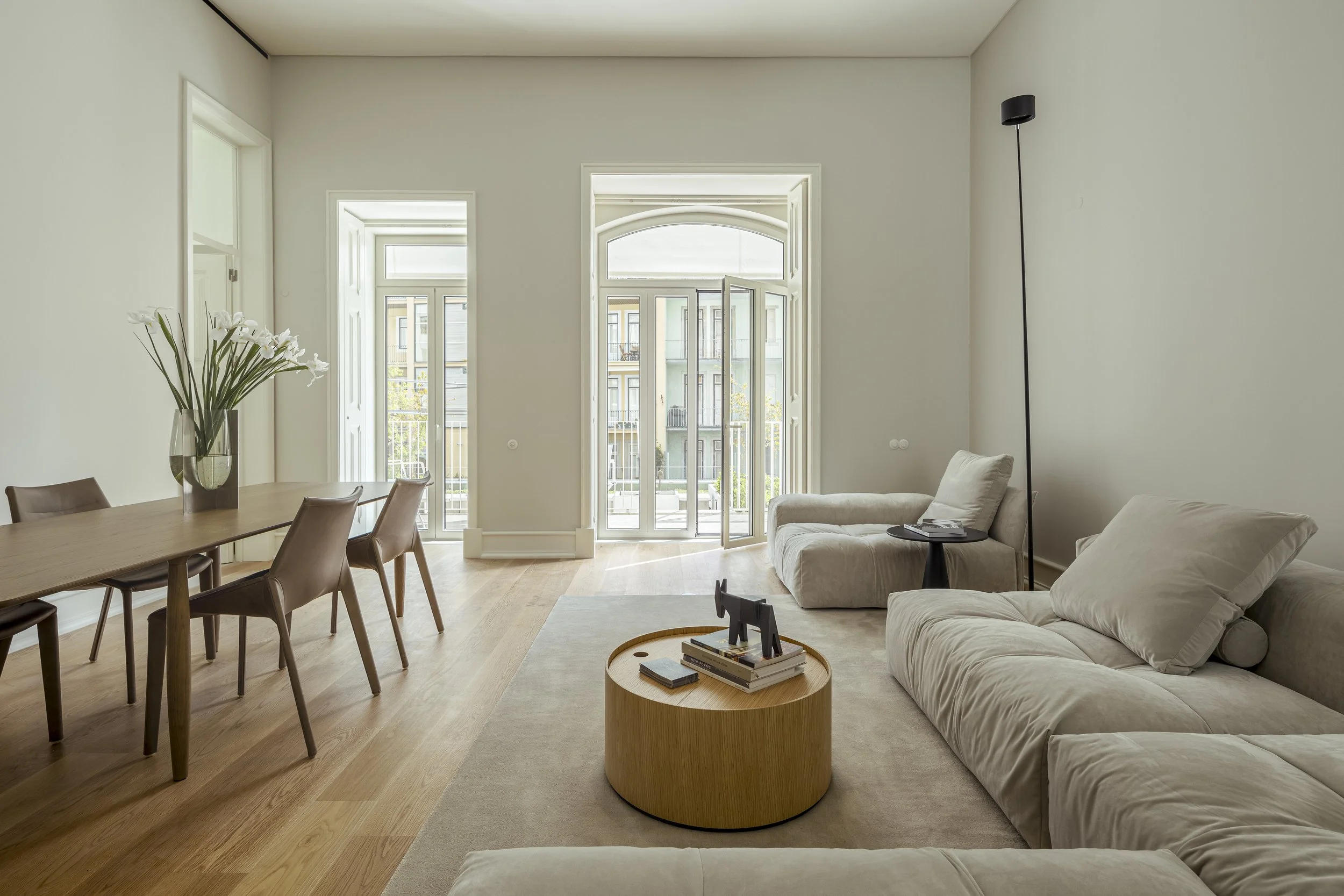 Bright living room with beige sofas, wooden coffee table, and large balcony doors with glass panels, white walls, and wooden flooring.